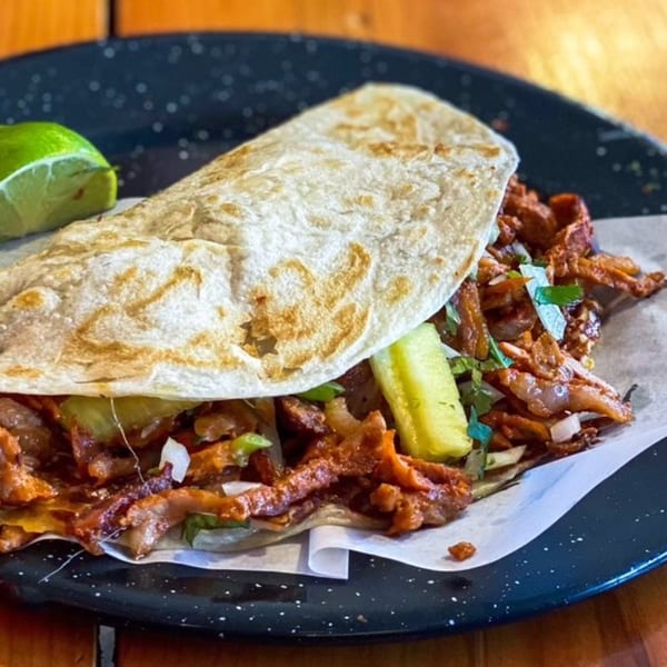 Al pastor taco from Holy Taco, with marinated pork, pineapple, onions, and cilantro, served with a lime wedge.