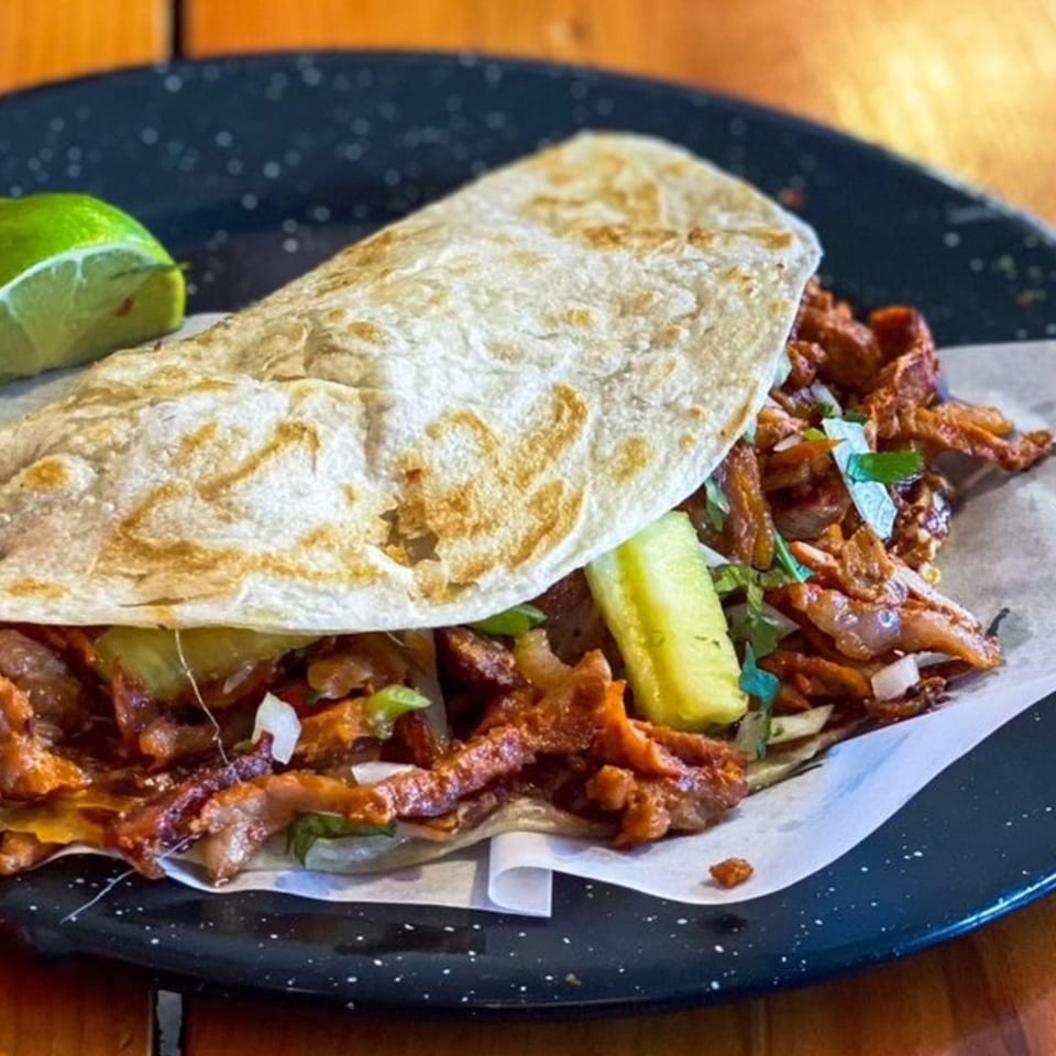 Al pastor taco from Holy Taco, with marinated pork, pineapple, onions, and cilantro, served with a lime wedge.