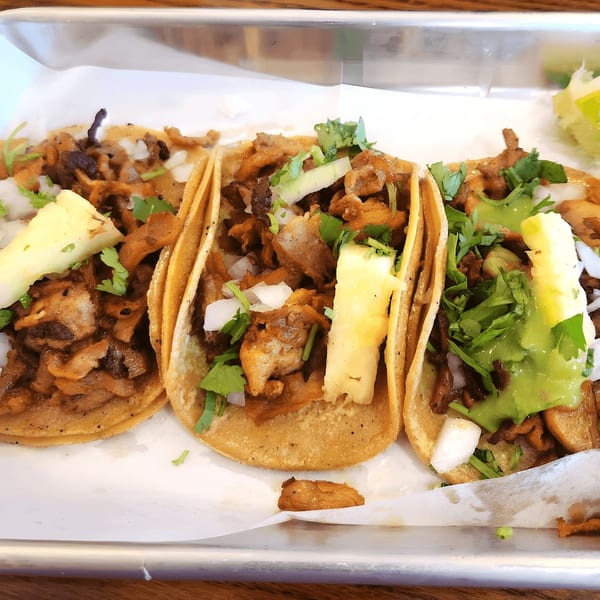 Three Holy Taco al pastor tacos with pineapple, onions, cilantro, and green sauce on a tray.