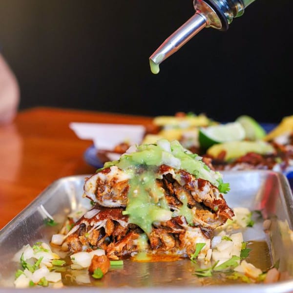 Holy Taco's tacos with green sauce being drizzled from a metal pour spout onto them.