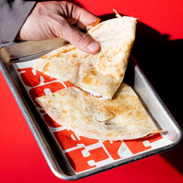 Hand picking up a Holy Taco quesadilla from a metal tray with red and white branded paper.