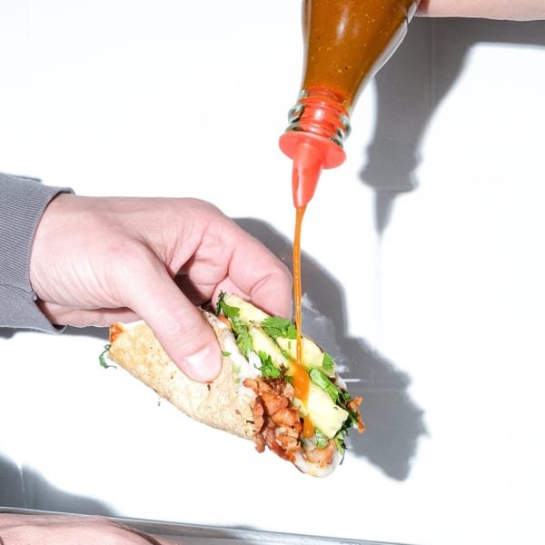 A hand holds a Holy Taco al pastor taco as hot sauce is squeezed onto it.