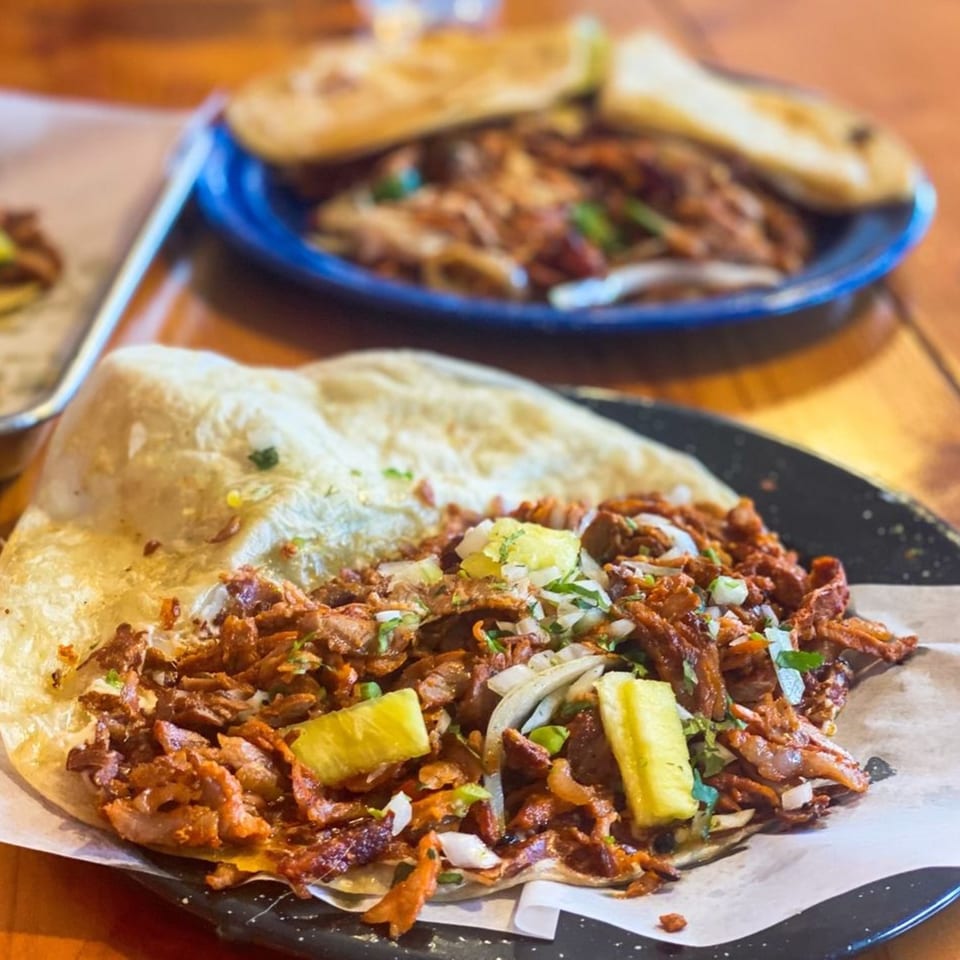 Al pastor taco with pineapple, onions, and cilantro on a black plate, with more tacos in the background.