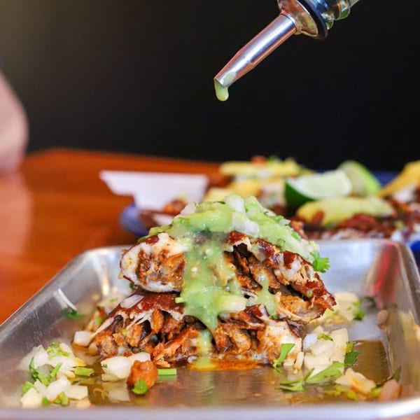 Holy Taco's tacos with green sauce being drizzled on them, served on a metal tray.