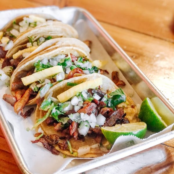 Four Holy Taco al pastor tacos with pineapple, onions, cilantro, and lime wedges on a metal tray.