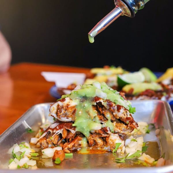 Holy Taco's tacos with green sauce being drizzled from a metal spout onto them.